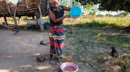 A woman in colourful dress pouring grain from a blue basket to a pink one, in a sparsely populated farm