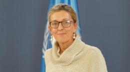 A woman in a white sweater and glasses standing in front of a blue flag with the UN logo on it.