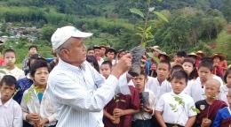 A man in a white shirt and white cap holds up a sapling in front of a group of school students.