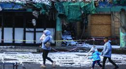 A woman walks in front of destroyed buildings and holds a child in her arms, with two children walking behind her.
