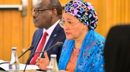 A woman in a blue dress and colourful headscarf, the UN Deputy Chief Amina J. Mohammed, speaks into a microphone at a podium. Another man in a blue suit and a red tie sits to her right side.