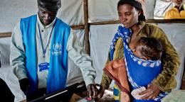A woman holds a baby in her arms while a man in a blue United Nations vest looks at a computer.
