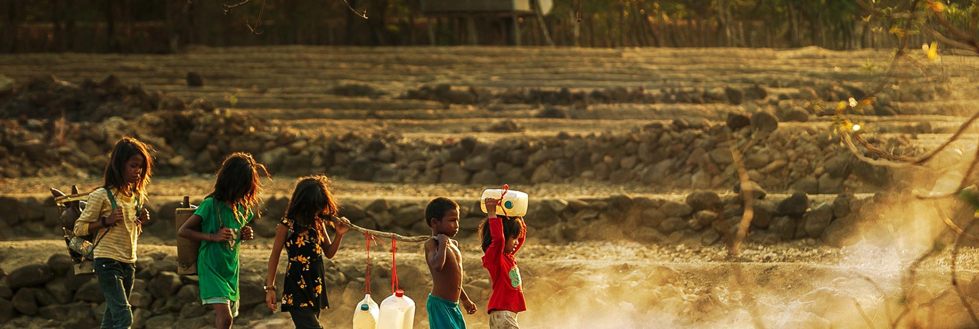 Little children walk in a line as they all hold a large stick holding jugs of water. 