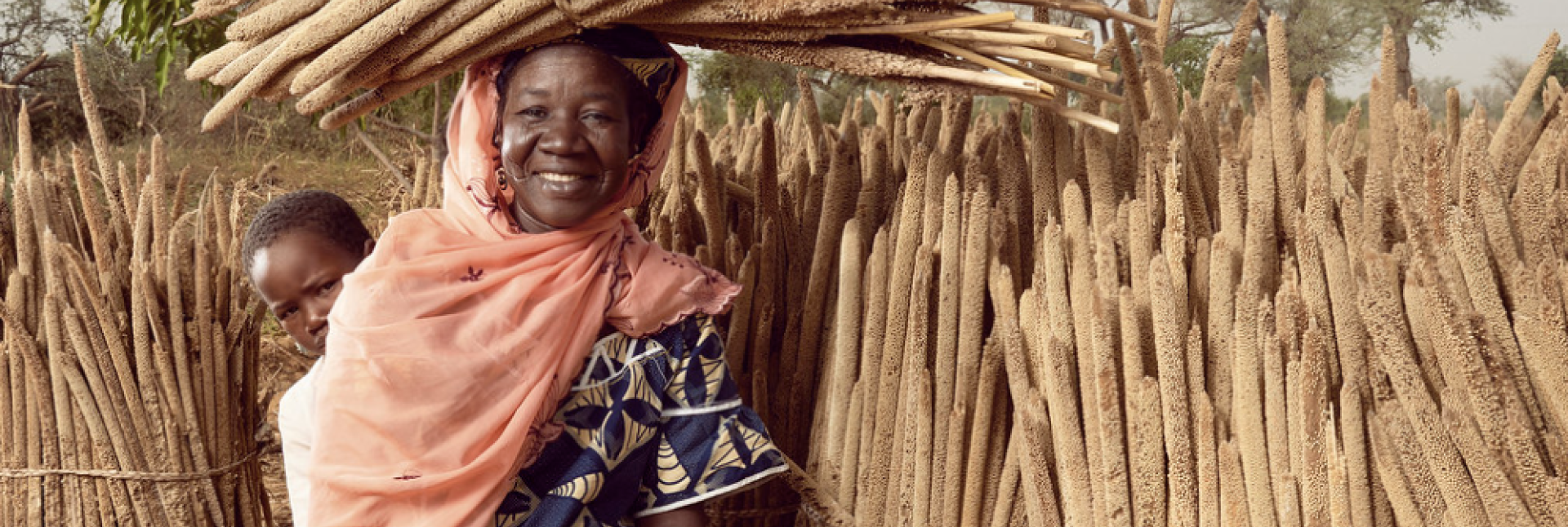 A smiling woman holding a baby in an orange sling sits while balancing wheat on her head. 