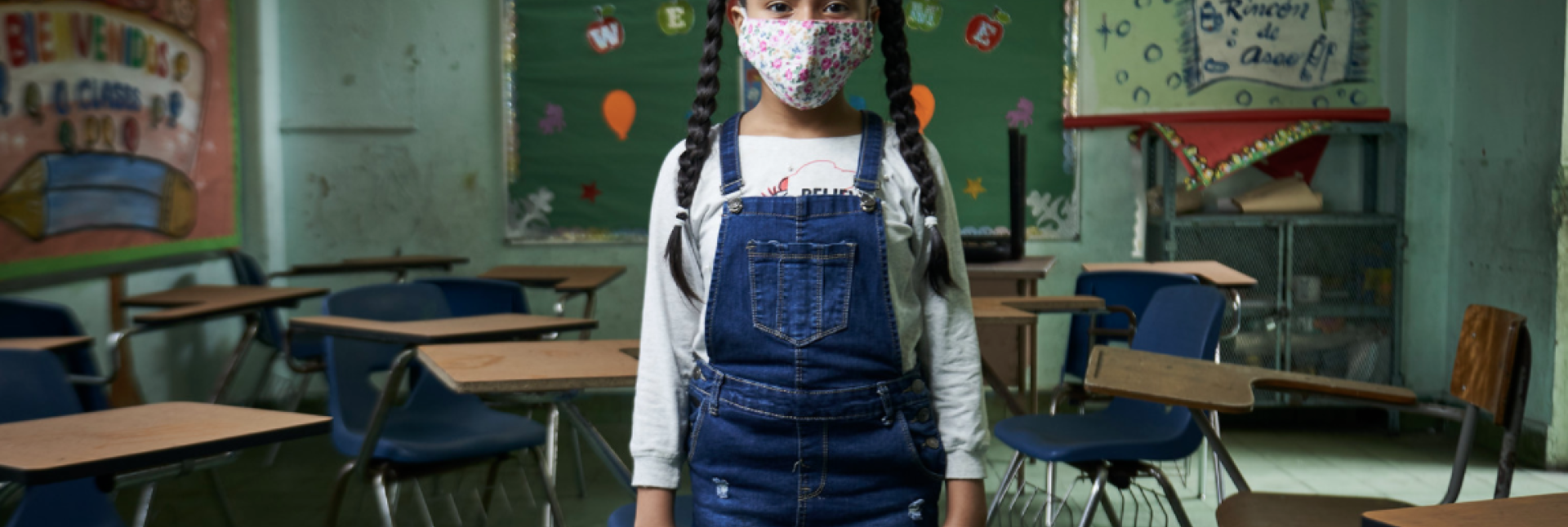 A young girl in a white face mask and blue overalls stands in a classroom. 