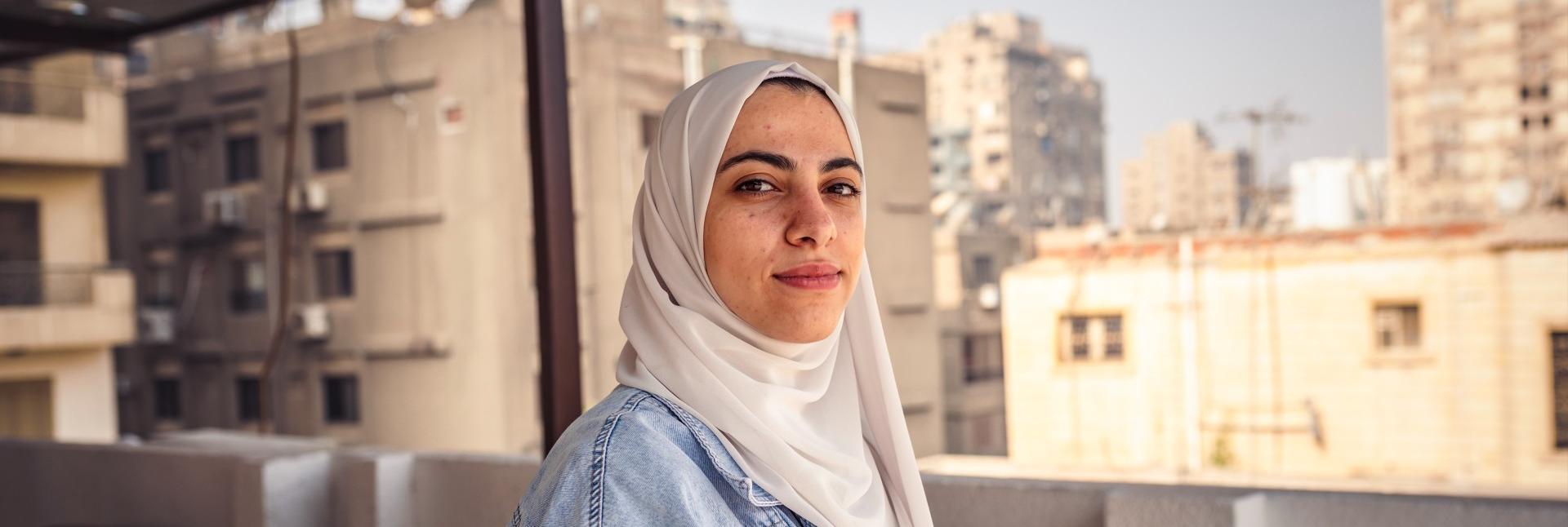 A girl in a white head scarf and denim jacket faces the camera, on a sunny rooftop amid tan-colored high-rise buildings. 