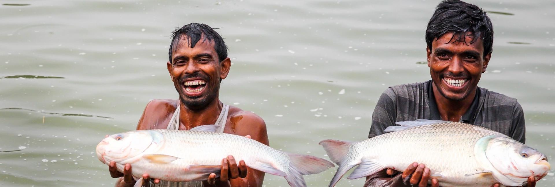 Two men hold big cream-colored fishes and laugh, while standing in water.