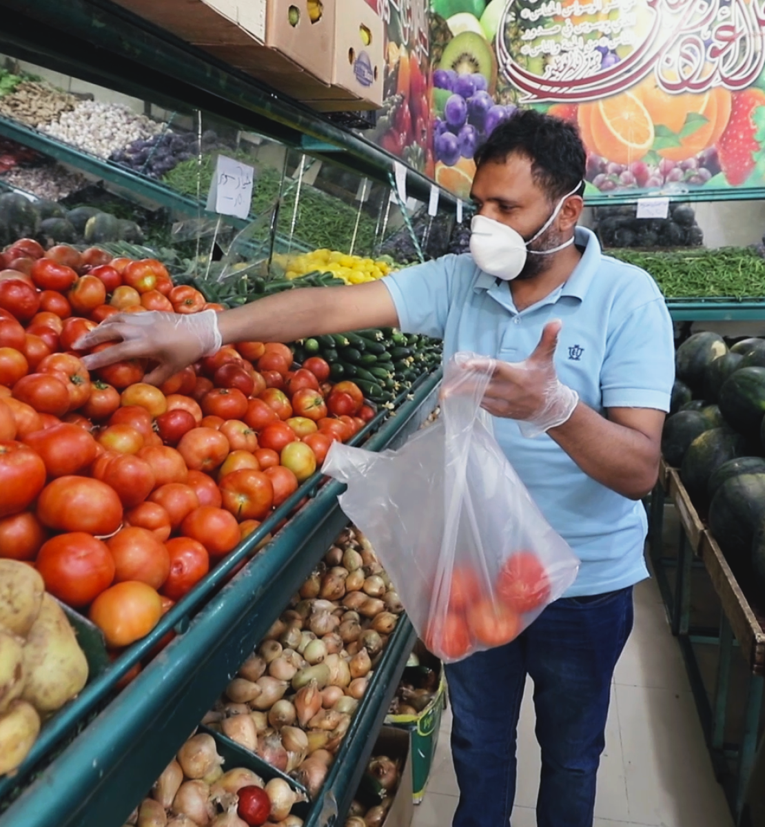 Abdou, un réfugié yéménite à Amman, fait ses courses chez un marchand de légumes après la réouverture des petits commerces. Il porte un masque de protection et des gants. Au premier plan de l’image, un enfant, lui aussi portant un masque, le regarde mettre des tomates un sac en plastique.