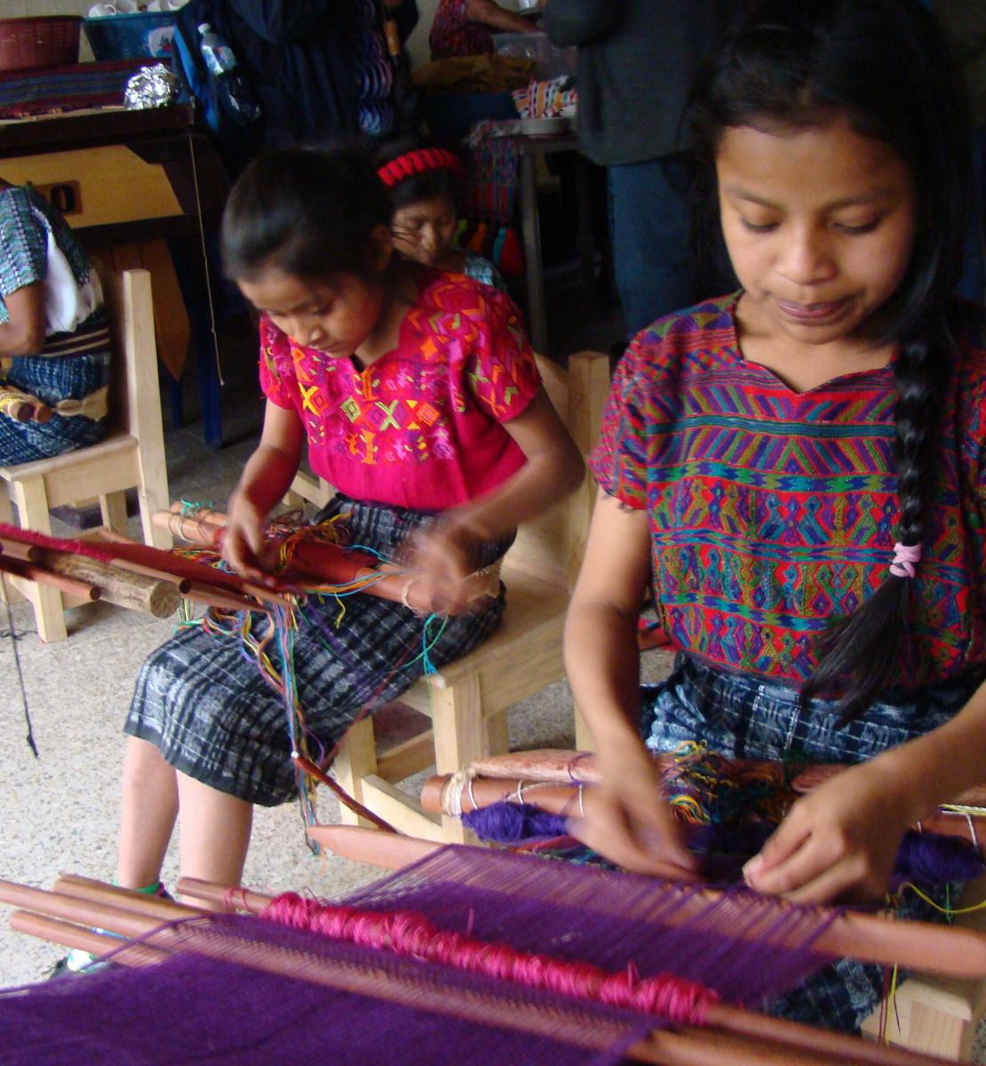 Photo showing three indigenous girls sitting on a chair and learning to weave using a traditional loom 