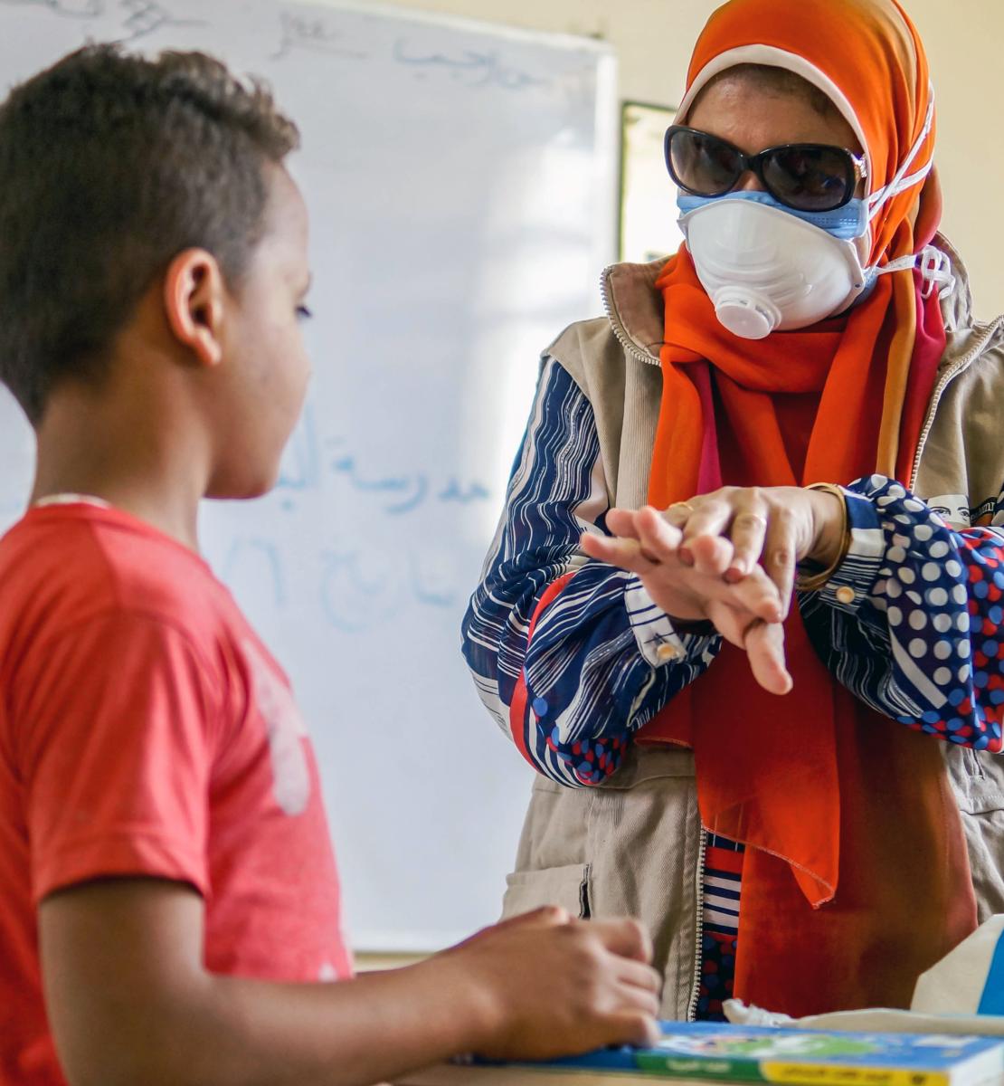 Un membre féminin du personnel de l'ONU portant un masque de protection donne à un jeune garçon des conseils de sécurité en lui montrant les gestes appropriés à faire pour un lavage efficace des mains.