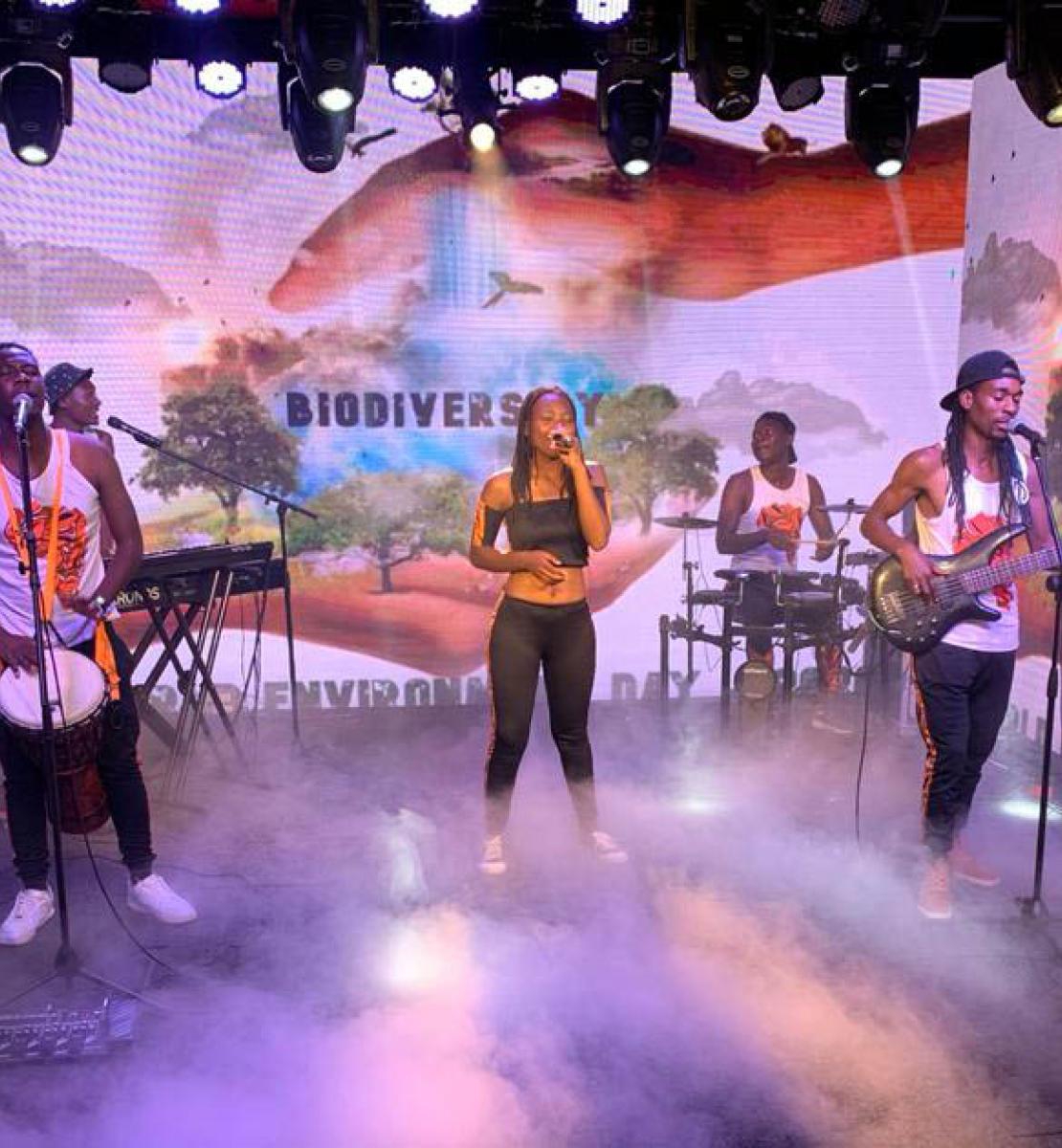 Caroleen Masawi and her band perform surrounded by green screens featuring images of the globe and the term "Biodiversity" featured on them.