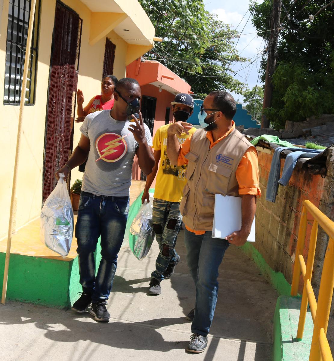 Residentes de la zona y personal de la ONU pasan frente a las casas de un barrio de la República Dominicana. Llevan una máscara protectora y conversan entre ellos mientras avanzan juntos.