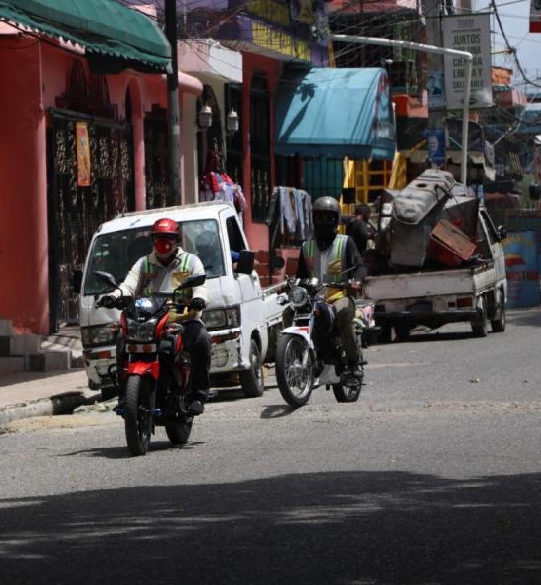 Escena de la vida citadina, donde se ve a motociclistas con protectores faciales y cascos de seguridad conduciendo por una calle; y varias camionetas estacionadas en los laterales de la calle.