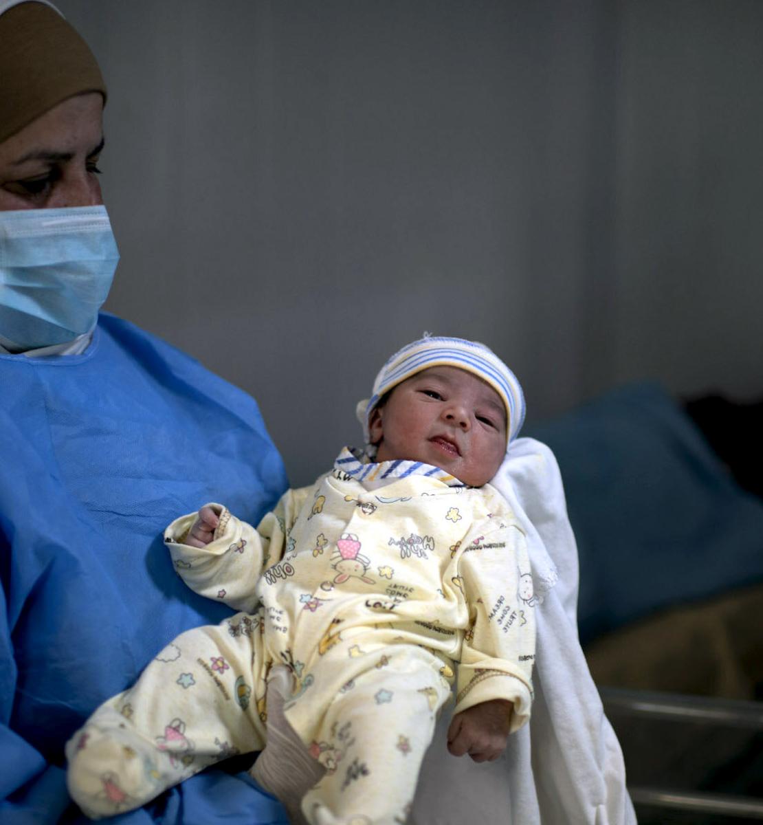 Une soignante portant un masque et une blouse de protection bleue tient un nouveau-né dans les bras.