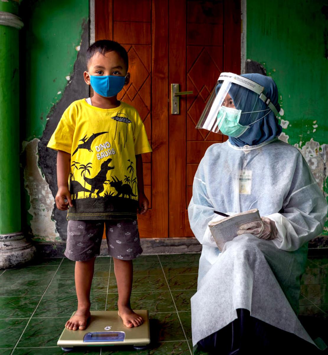 A little boy stands on a scale as he looks at the camera wearing a face mask and a healthcare worker in full head-to-toe PPE kneels by him.