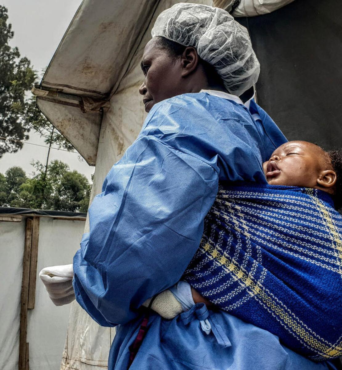 A woman wearing personal protective equipment carries a small baby on her back. 
