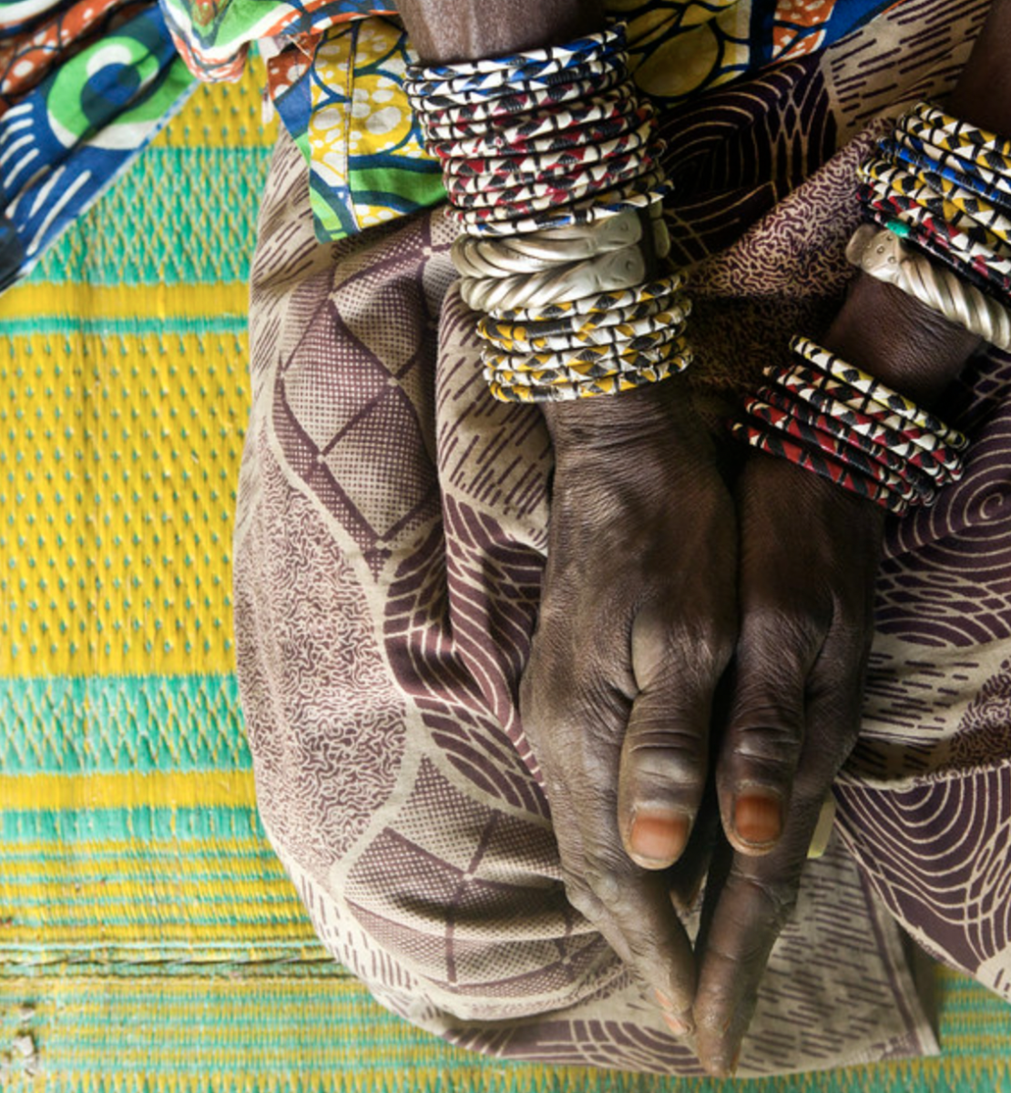 A woman's hands, with several bracelets, rest on her knees on top of a bright colored cloth.