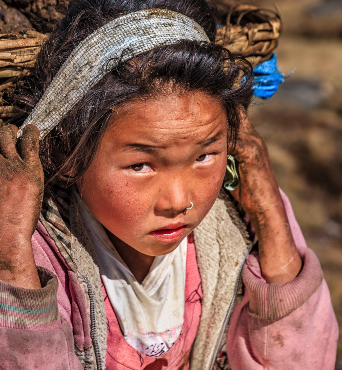 A young girl, with dirt on her hands and face, carries a large basket on her back.