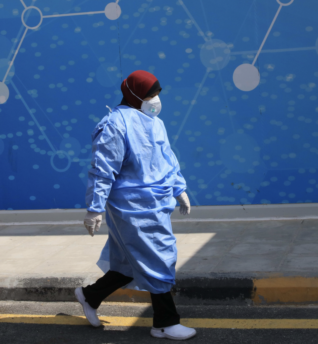 Una enfermera con equipo médico completo y una mascarilla camina por la calle cerca de un edificio azul. 