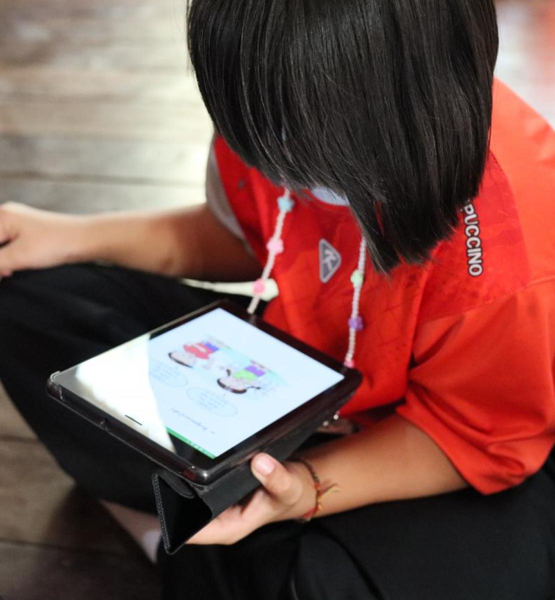 Un enfant vêtu d'un t-shirt orange foncé et portant une coupe de cheveux au carré est assis sur le sol en tailleur et regarde des dessins sur une tablette.