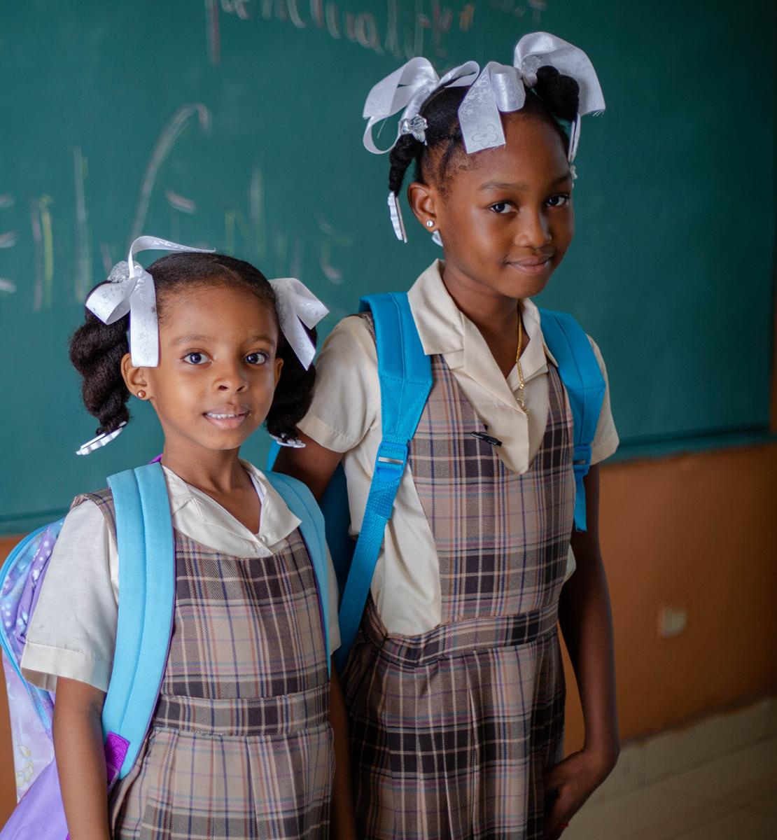 Dos niñas con mochila y uniforme escolar se sitúan frente a una pizarra.