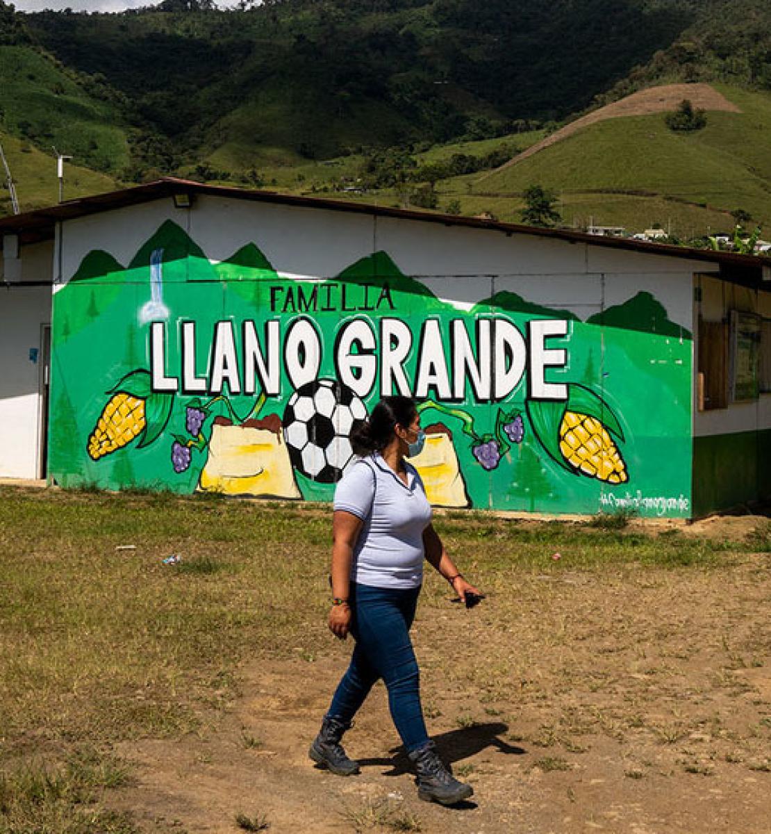 A woman wearing a face mask walks in front of a house with a painted green mural of food and a soccer ball. 