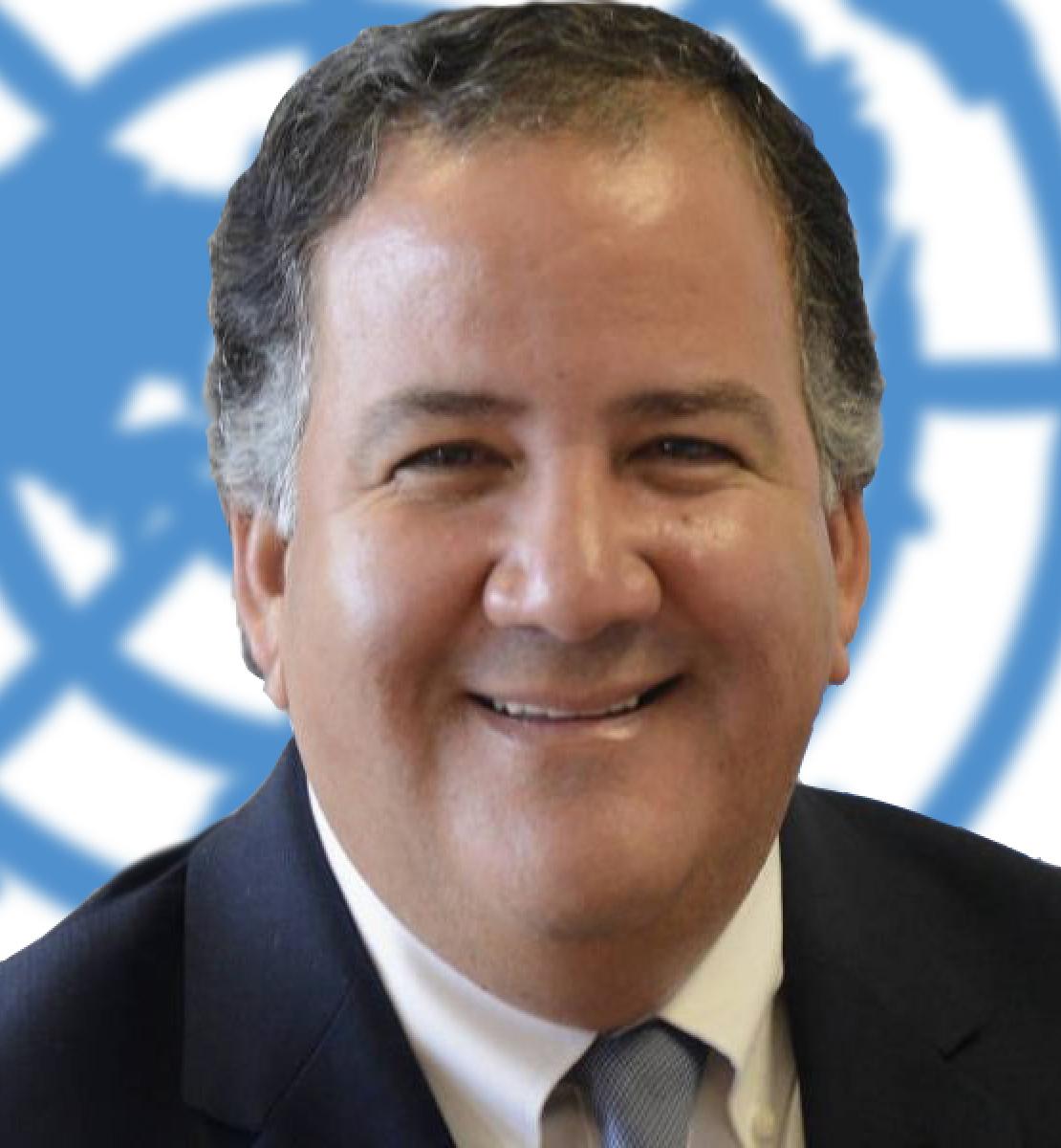 Un hombre sonriente mira directamente a la cámara con el logotipo de las Naciones Unidas de fondo.