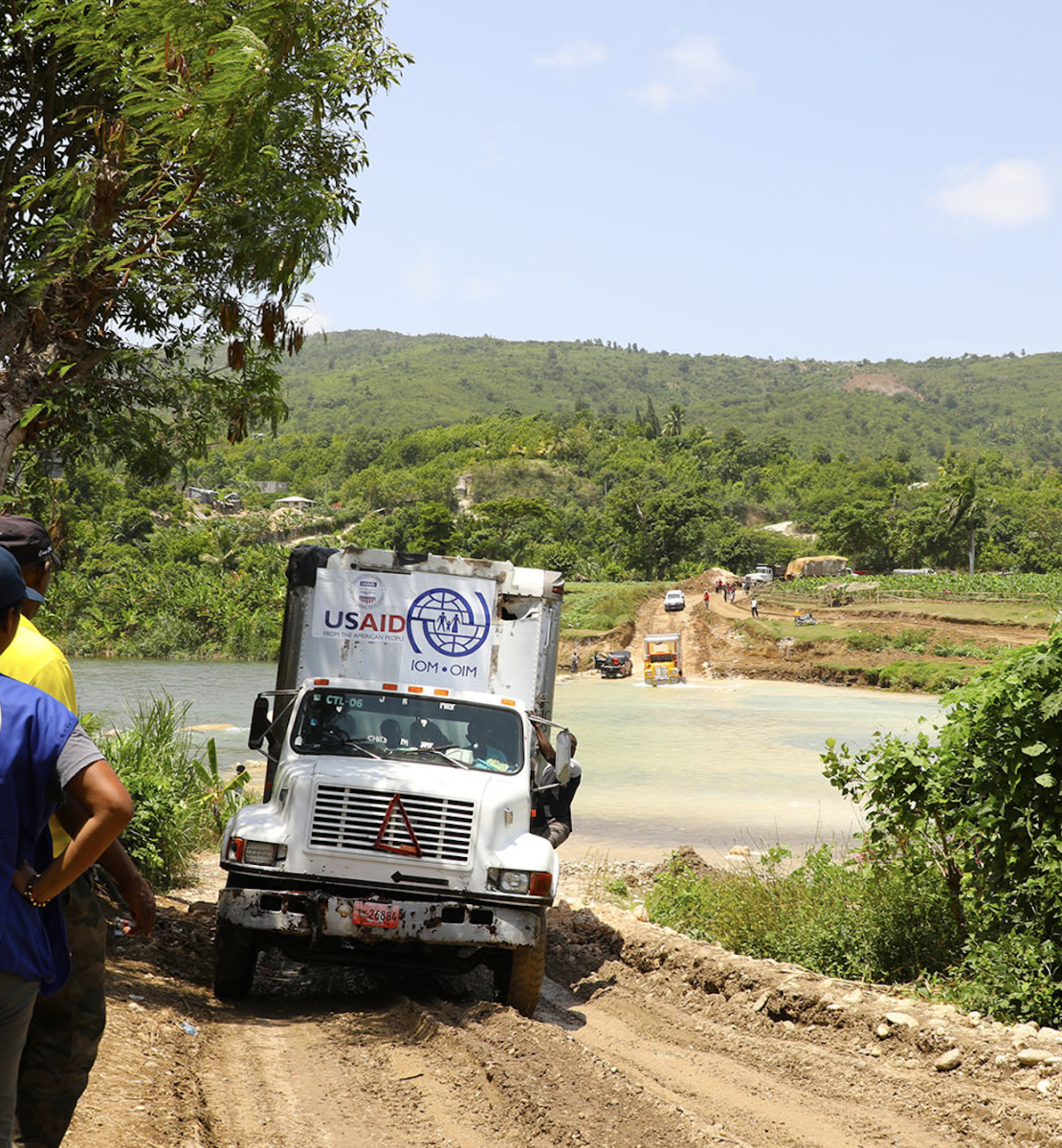 Los trabajadores de la Organización Internacional para las Migraciones se sitúan a un lado de la carretera mientras un gran camión circula por el camino de tierra y los niños y niñas miran desde arriba de un montículo.