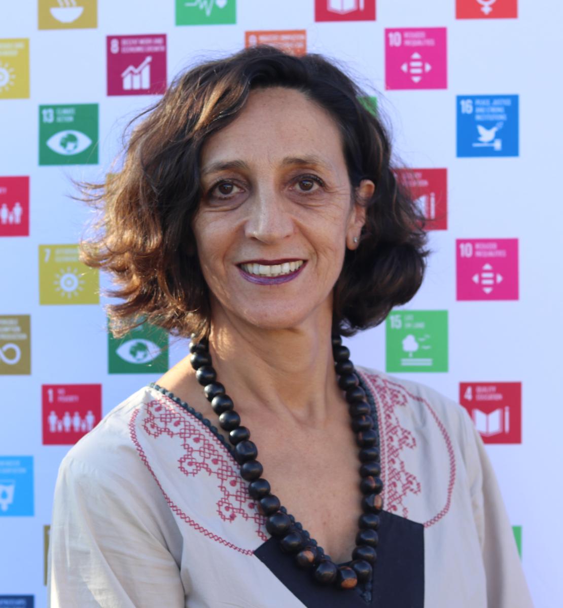 امرأة مبتسمة تقف أمام ملصق لأهداف الأمم المتحدة في مجال التنمية المستدامة.