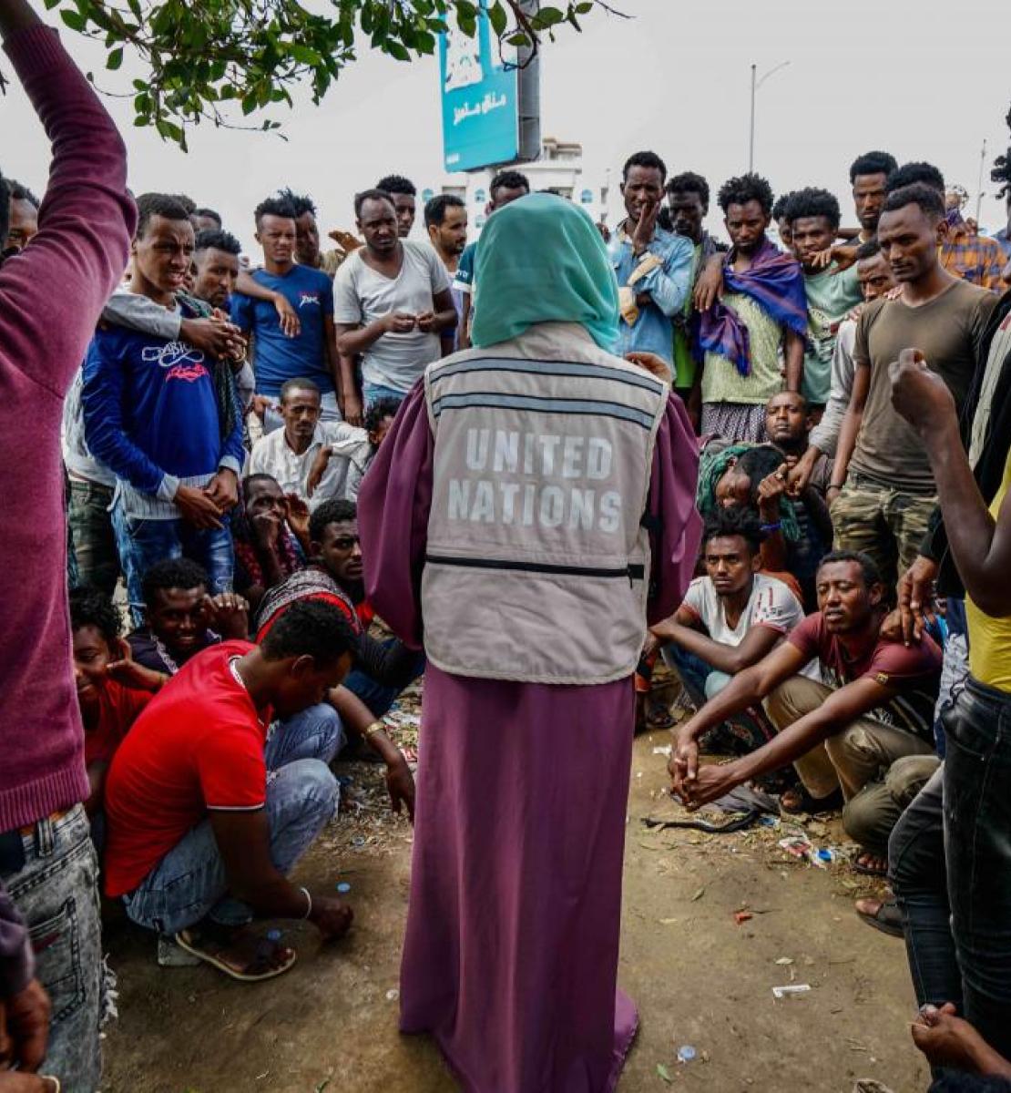 A woman in a United Nations vest is surrounded by many people waiting to receive humanitarian assistance.