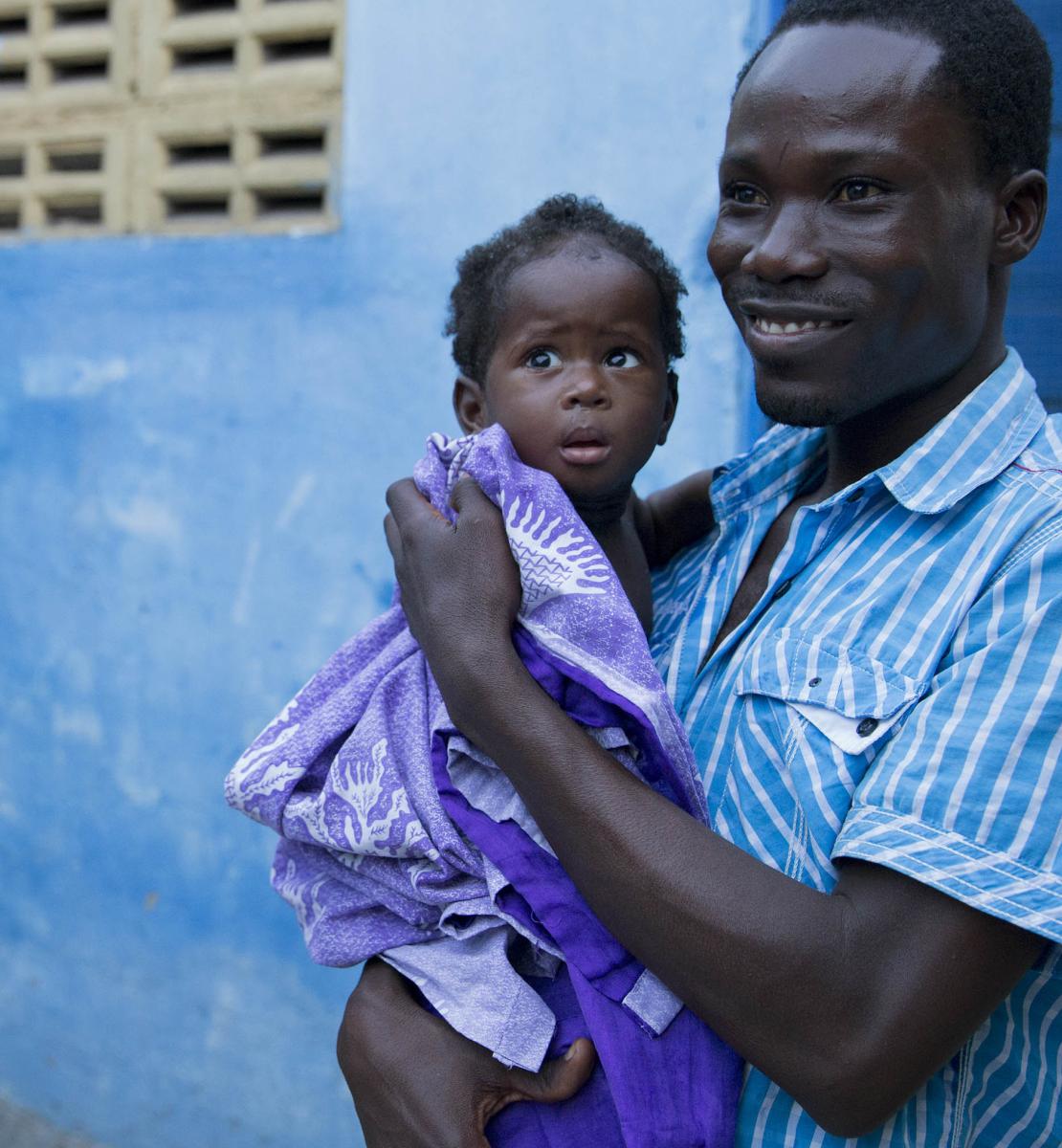 A father proudly holds his baby as stands in front of a building. 