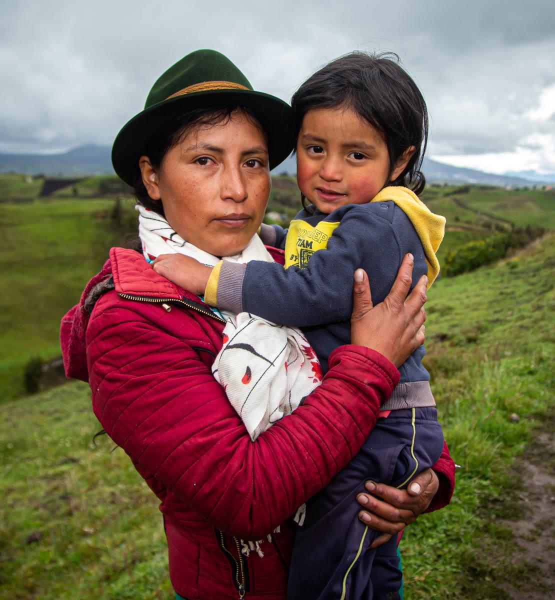 Une femme autochtone portant un chapeau se tient debout au milieu d'un champ, un jeune enfant dans les bras, par une journée nuageuse.