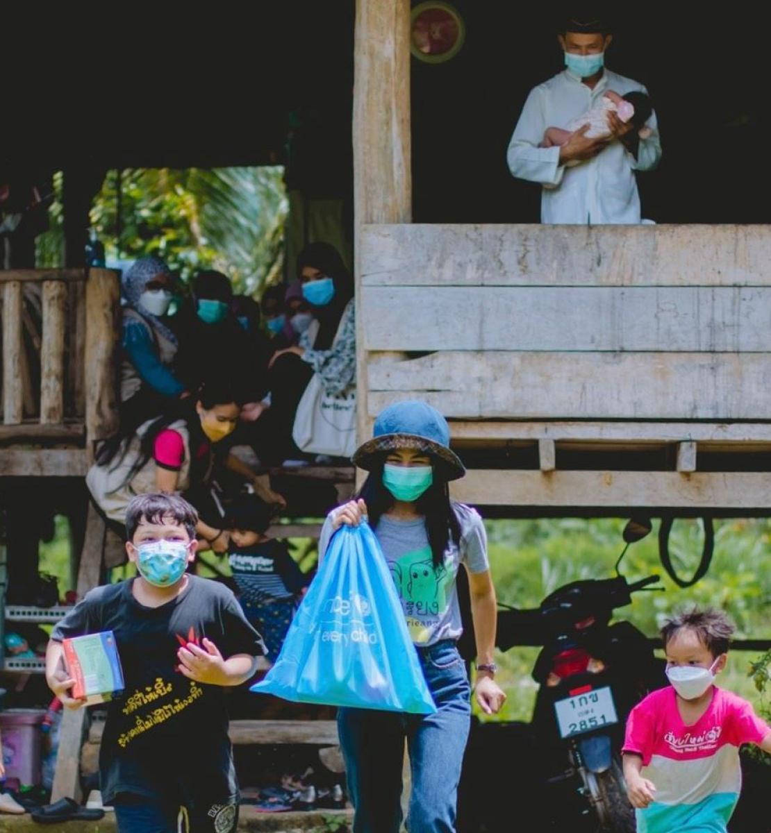 Les membres d'une famille, tous équipés d'un masque de protection respiratoire, sont assis sur la terrasse d'une maison en bois, tandis que plusieurs enfants portant également un masque marchent en direction de l'objectif en s'éloignent de la maison, divers objets distribués par l'UNICEF à la main.