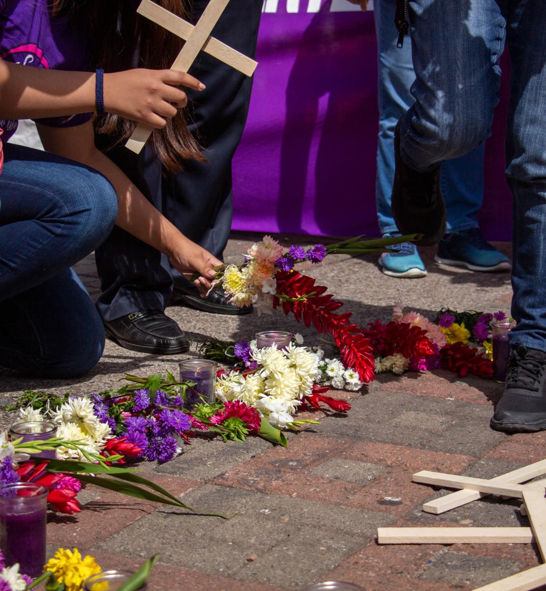 La imagen muestra los brazos de las personas que sostienen cruces arrodilladas junto a flores y más cruces amontonadas.