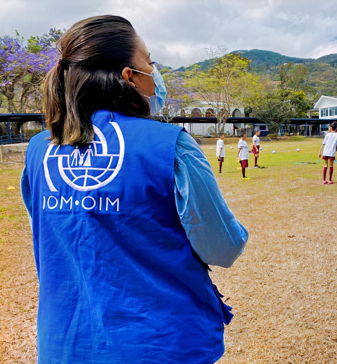 Una mujer con un chaleco de la OIM mira hacia un campo de fútbol con muchos niños y niñas jugando al fútbol.