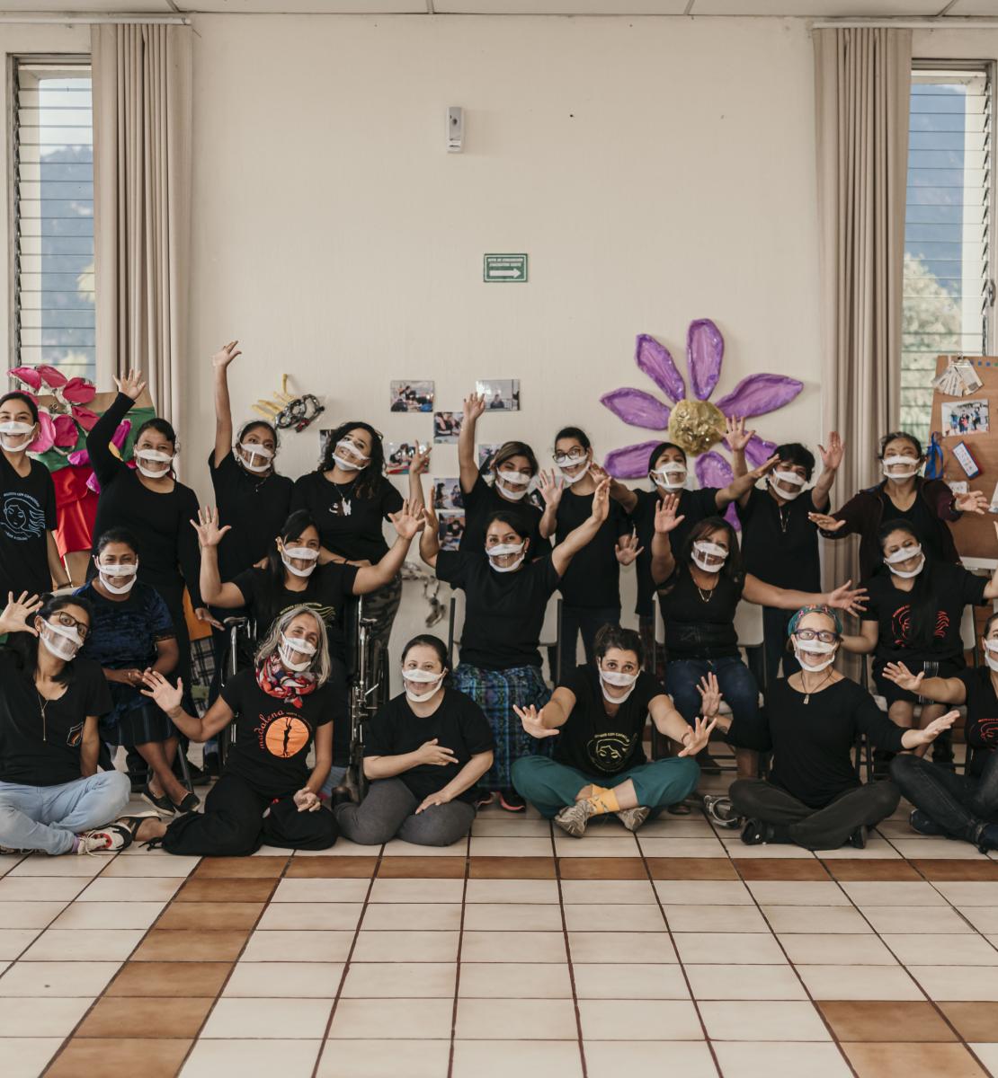 Members of the organization "Mujeres con Capacidad de Soñar a Colores" pose together against a wall as they cheerfully spread their arms towards the front.