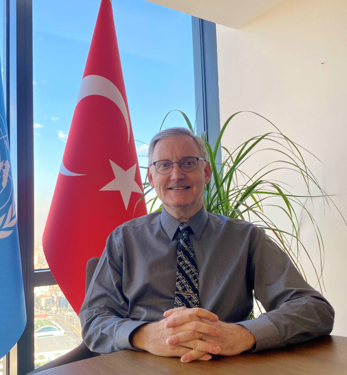 新任联合国驻土耳其协调员阿尔瓦罗·罗德里格斯的官方照片。他坐在办公桌前，双手合十，背对联合国和土耳其的旗帜微笑。