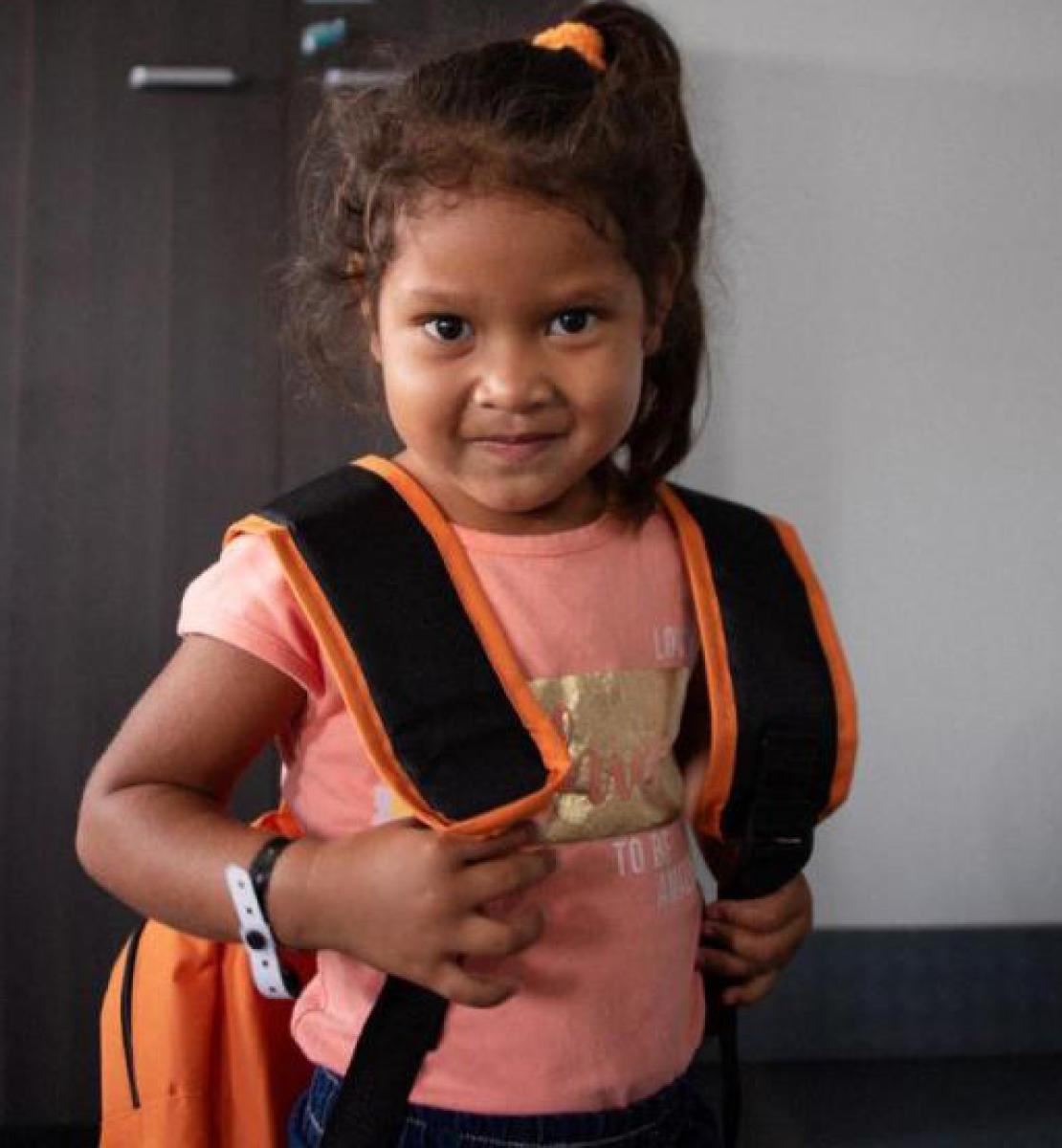A little girl with a small smile and looks stoically towards the camera as she grasps the straps of her backpack.