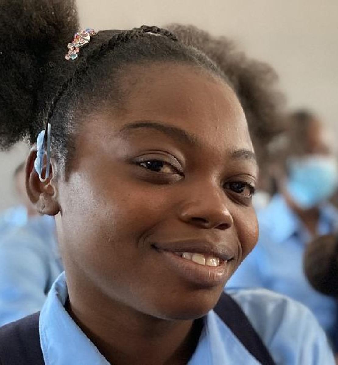 Una niña con su uniforme escolar sonríe a la cámara mientras el resto de los estudiantes miran hacia el centro del aula.