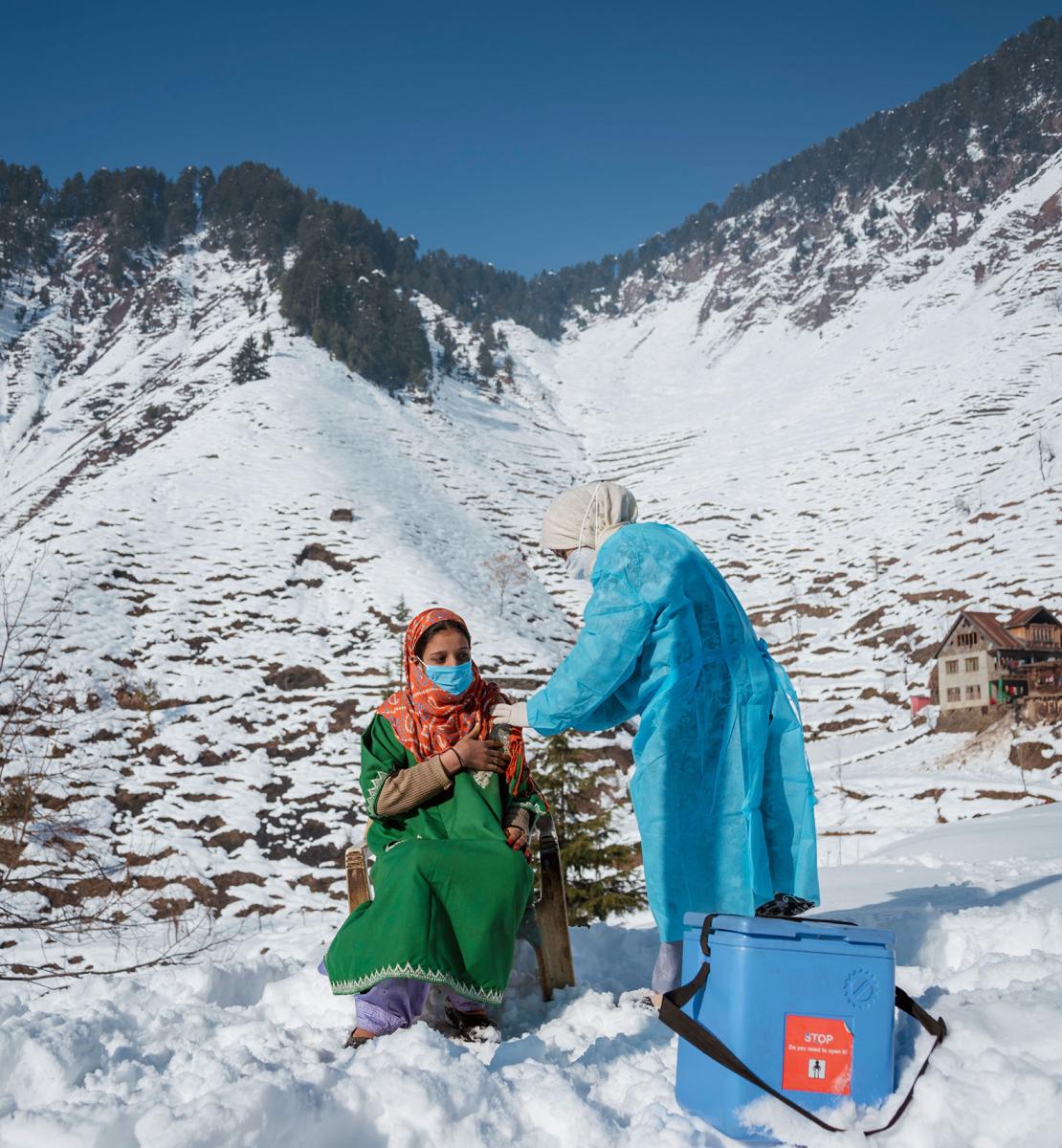 Une femme vêtue d’une robe verte se fait vacciner contre la COVID-19, près d’une glacière, par une agente de santé vêtue d’une blouse de protection bleue, au sommet d'une montagne enneigée, en Inde.
