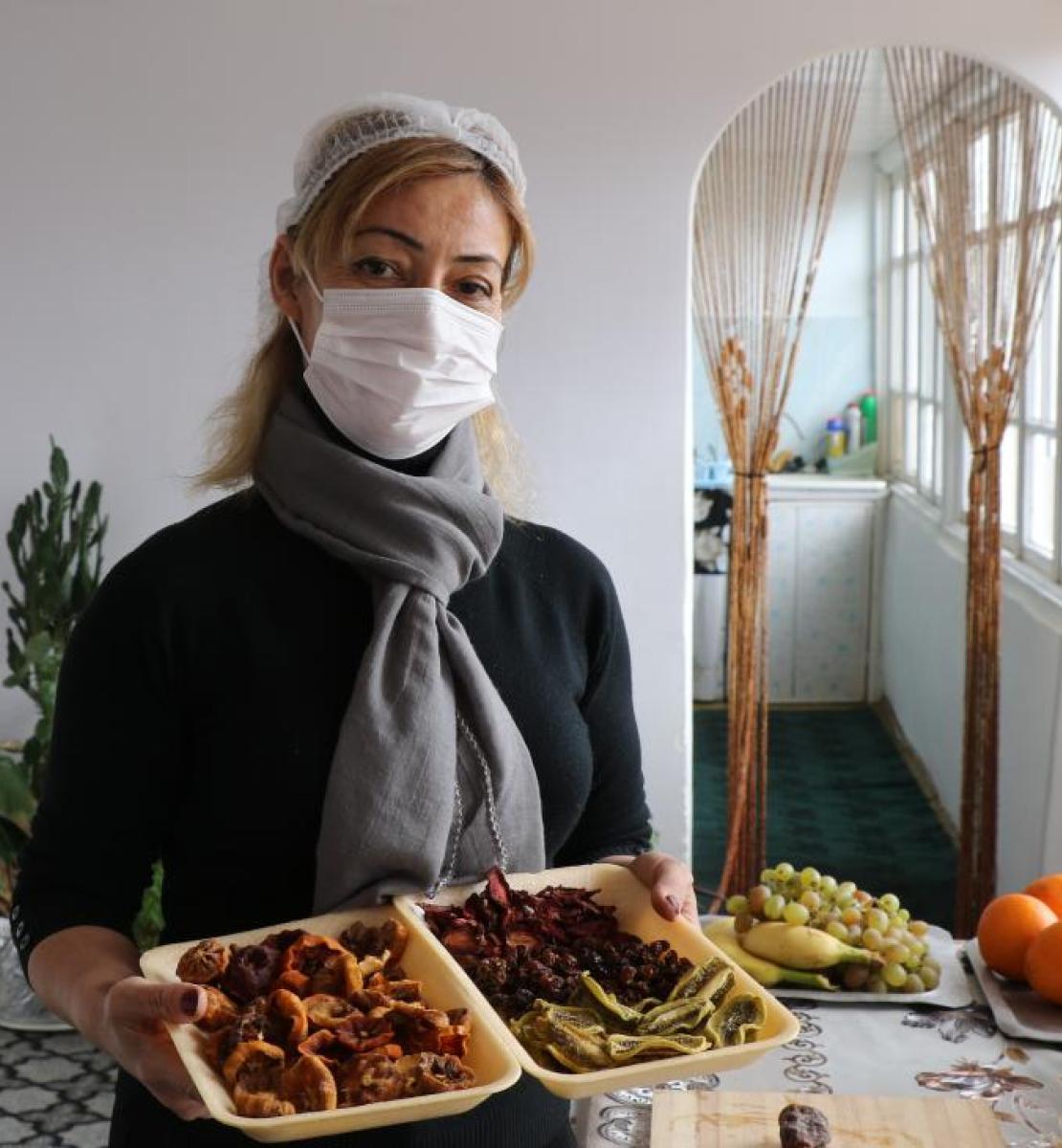 A woman wearing a white facemask showcases a takeaway food container full of dried fruits.