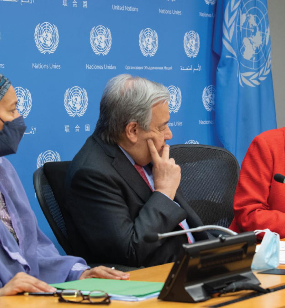 Un hombre, el Secretario General de la ONU, se sienta entre dos mujeres en medio de una animada discusión. Se aprecia una bandera la ONU y el logotipo de la ONU detrás de los presentes.