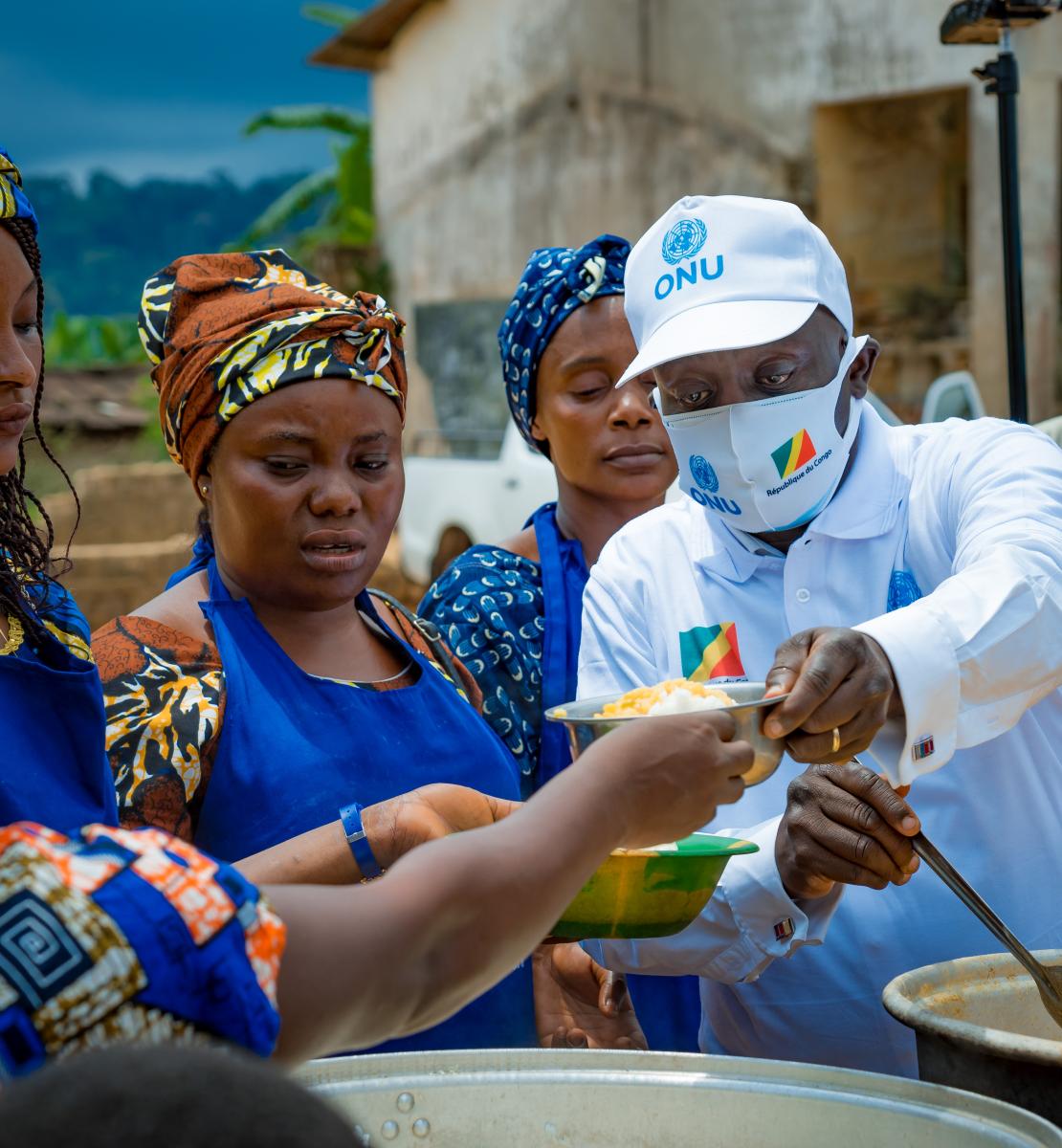 A man wearing a cap hands out food to women in blue and colored dresses. 