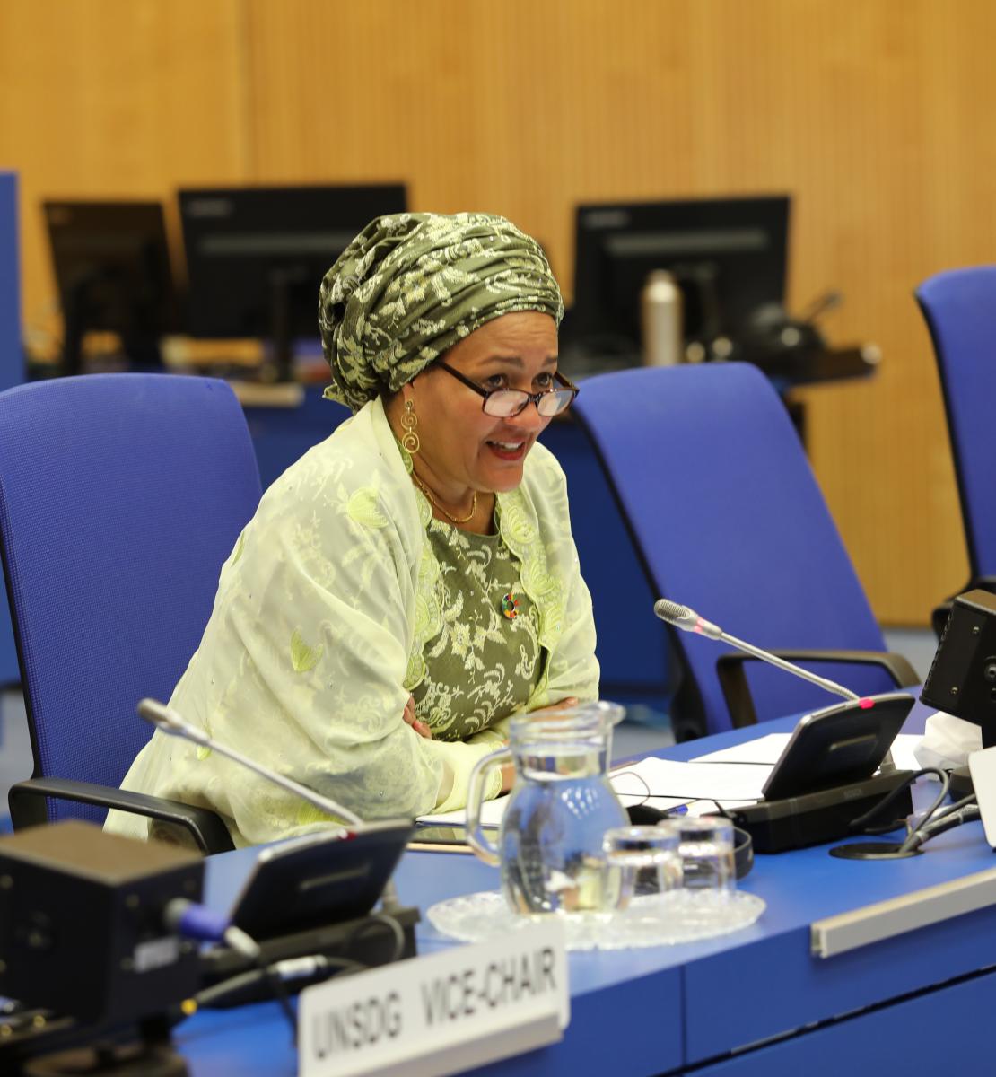 Une femme portant un châle jaune, un turban et des lunettes est assise à une table rectangulaire bleue, dans une salle de conférence, et parle dans un microphone lors d'une réunion organisée par l'ONU.
