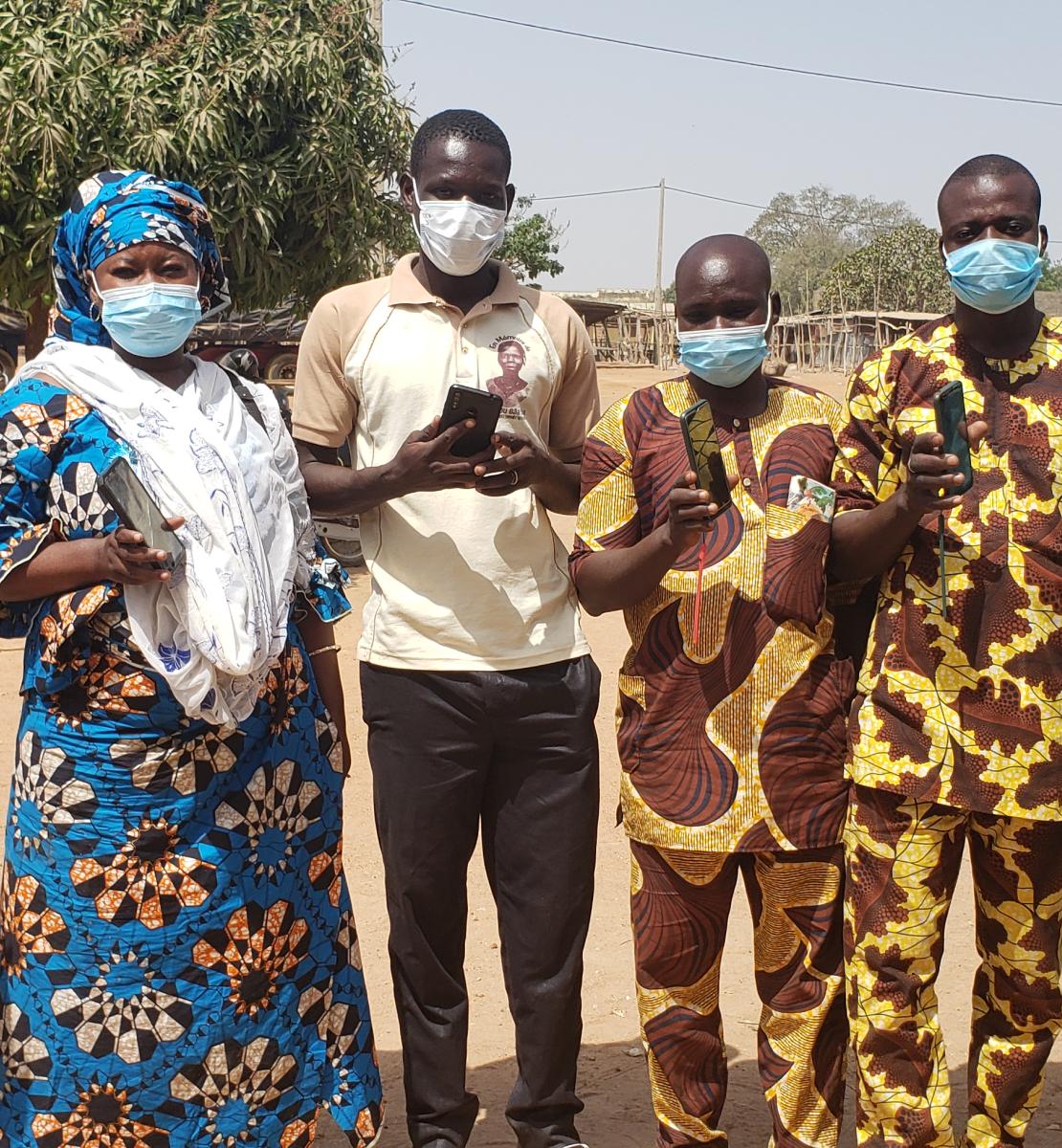 Un groupe de personnes portant un masque de protection respiratoire se tiennent debout, en extérieur, et montrent leur téléphone portable à la caméra.