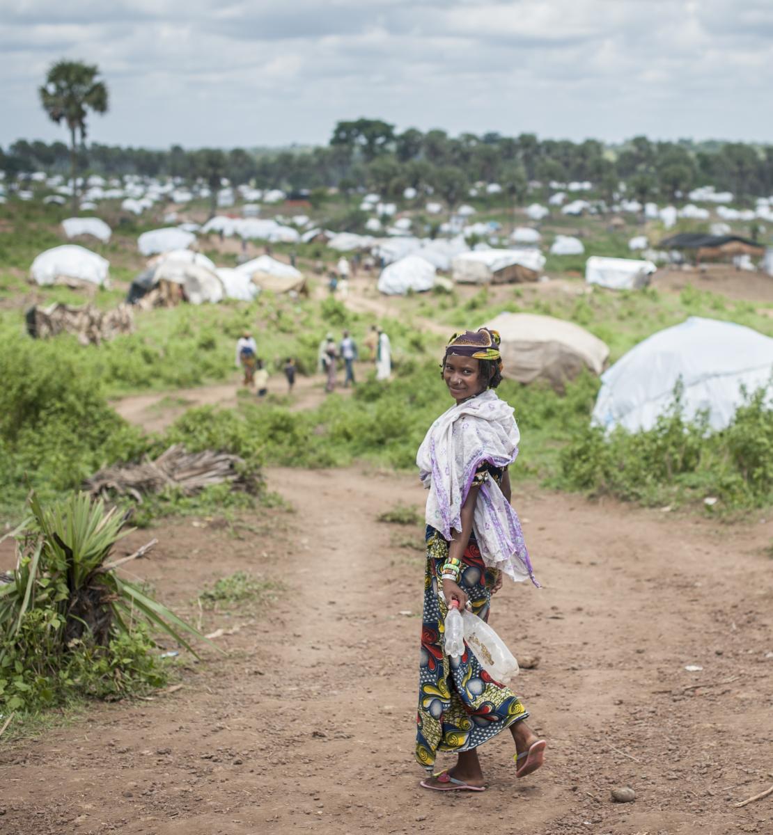 A woman walks toward a green field, dotted with tents, along a dirt road, and turns around to smile.
