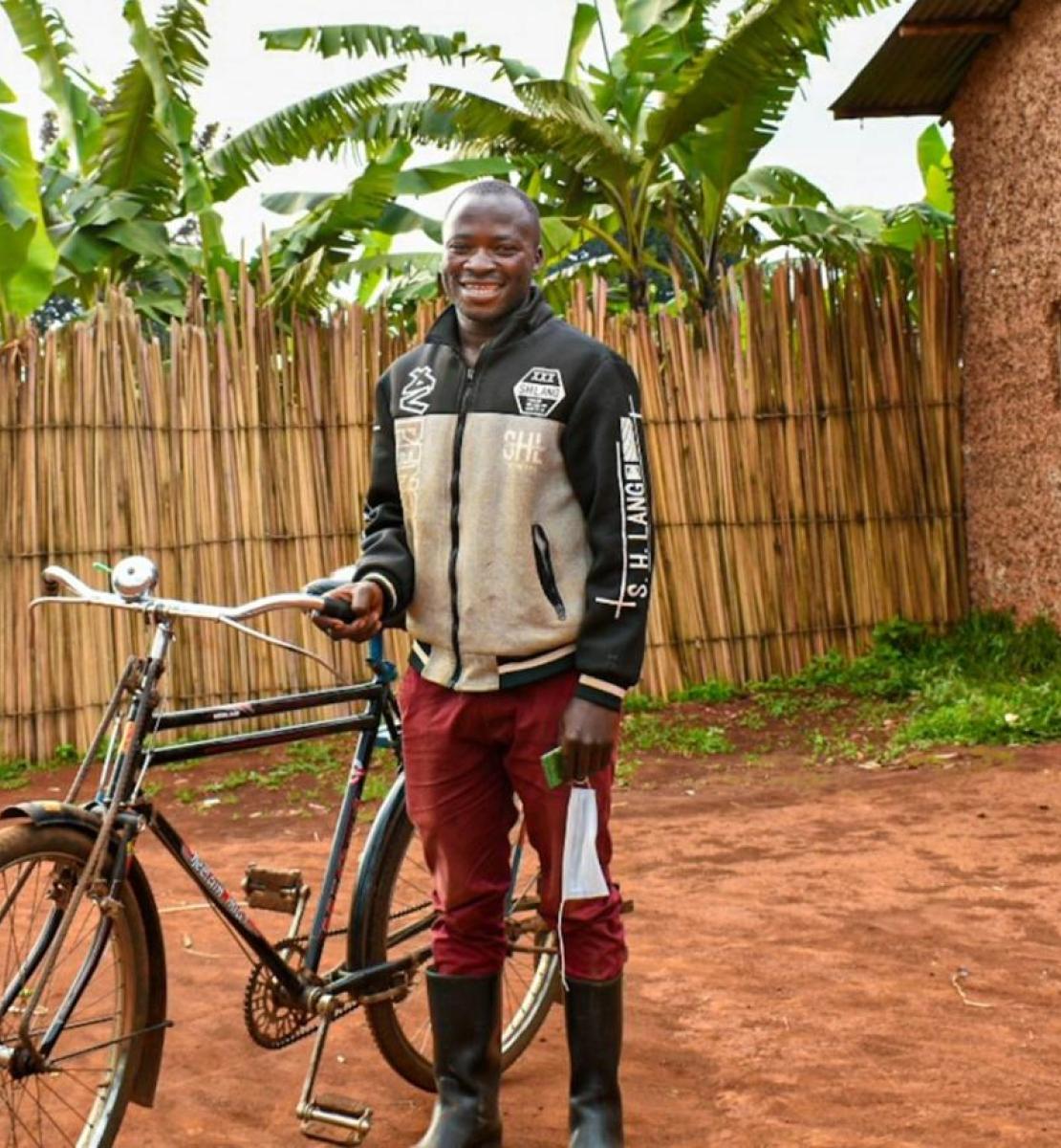 A man dressed in red pants stands by his bicycle outside a modest house lined with reeds and palm trees and smiles at the camera