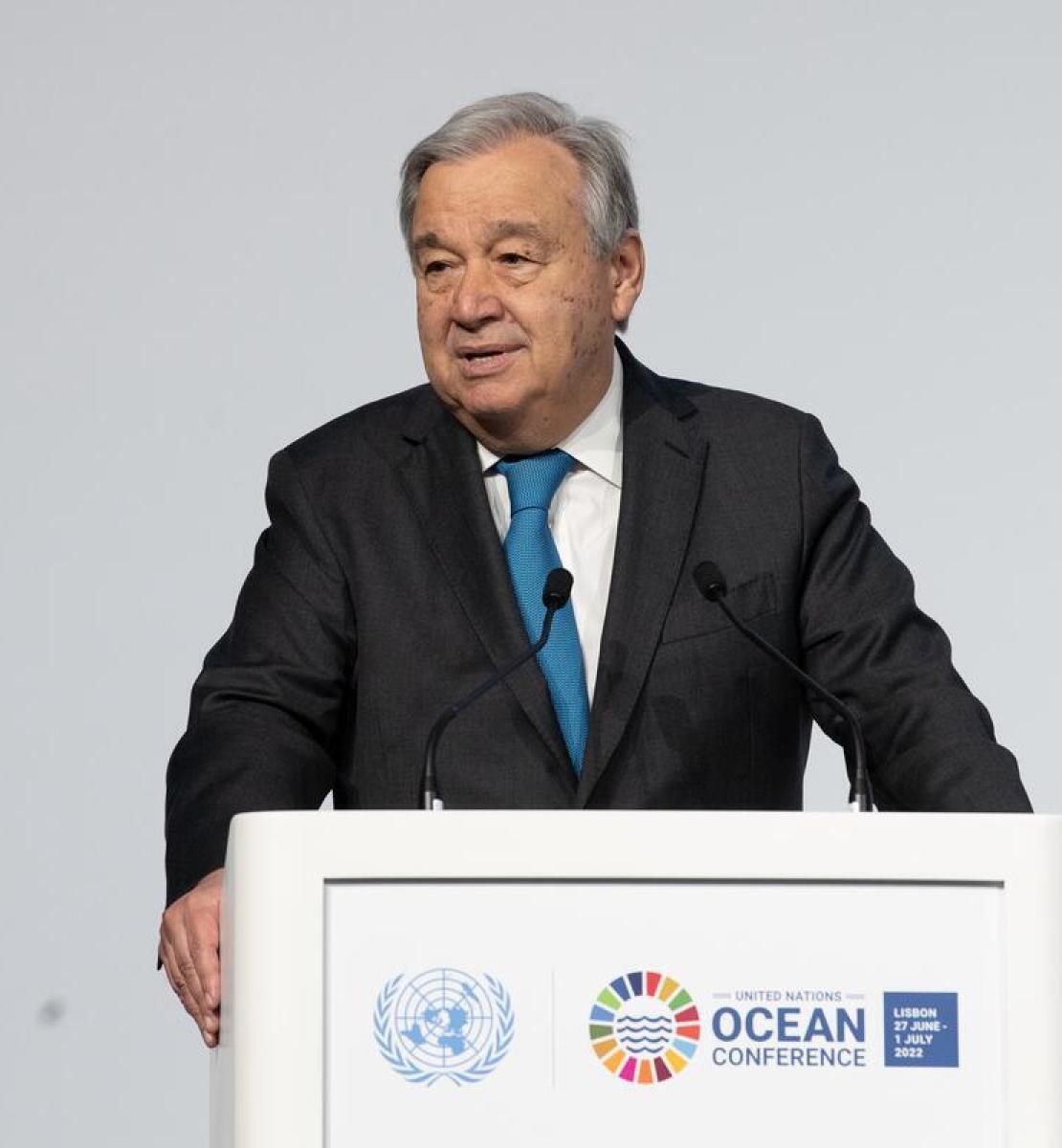 A man in a suit speaks into a microphone and a podium with UN logos on it, against a grey wall. 