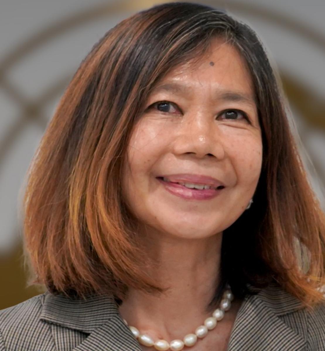 Une femme portant un collier de perles et un chemisier beige se tient face à la caméra en souriant, le logo de l'ONU en arrière-plan.