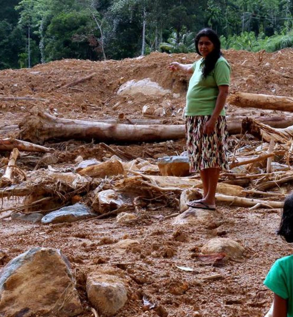 A woman in a mid-length skirt and t-shirt stands in a field of mud with tree trunk strewn around, talking to a young child.